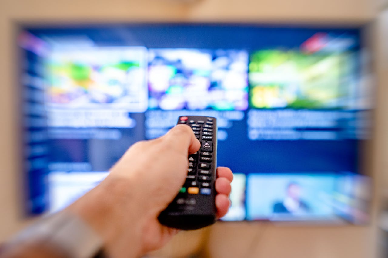 services-04 Hand holding remote control pointing at a blurred TV screen in a cozy indoor setting.